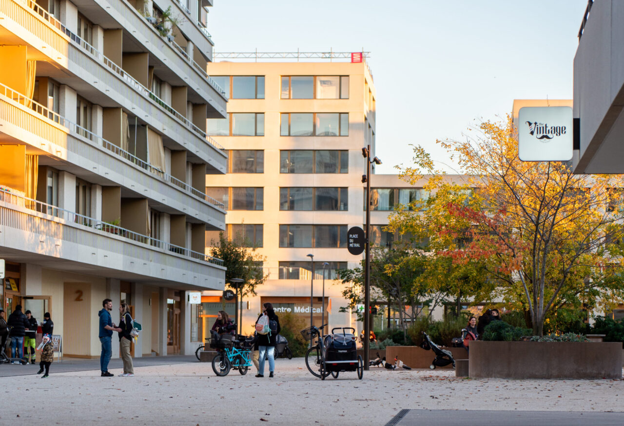 Place du Métral animée en automne, entourée d'immeubles contemporains, avec commerces et centre médical au rez-de-chaussée.