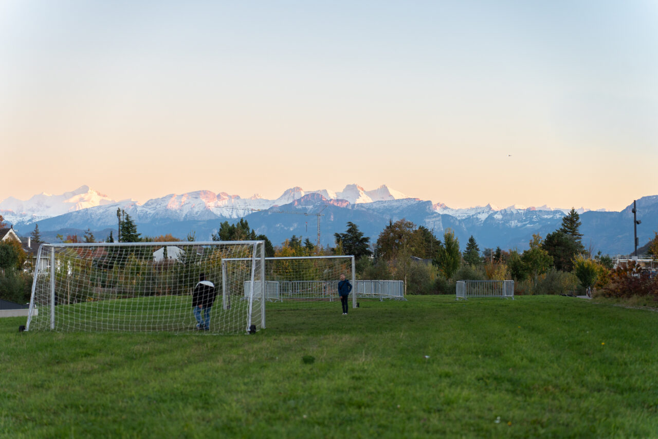 Terrain de football gazonné au coucher du soleil, avec vue sur les Alpes enneigées en arrière-plan.