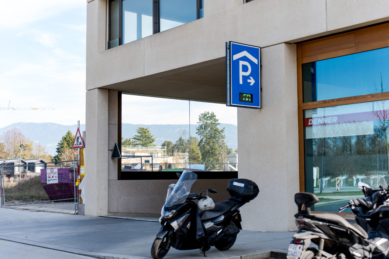 Panneau de signalisation d'un parking couvert avec compteur de places disponibles, en façade d'un immeuble mixte (commerces en rez-de-chaussée).